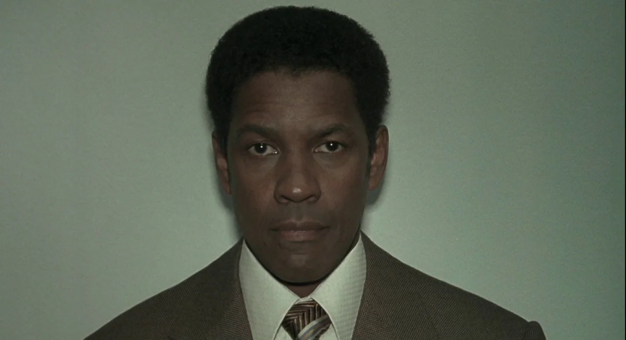 American Gangster (2007) movie still: closeup — A man stands against a plain, light green wall typical of a government office. The subject faces the…