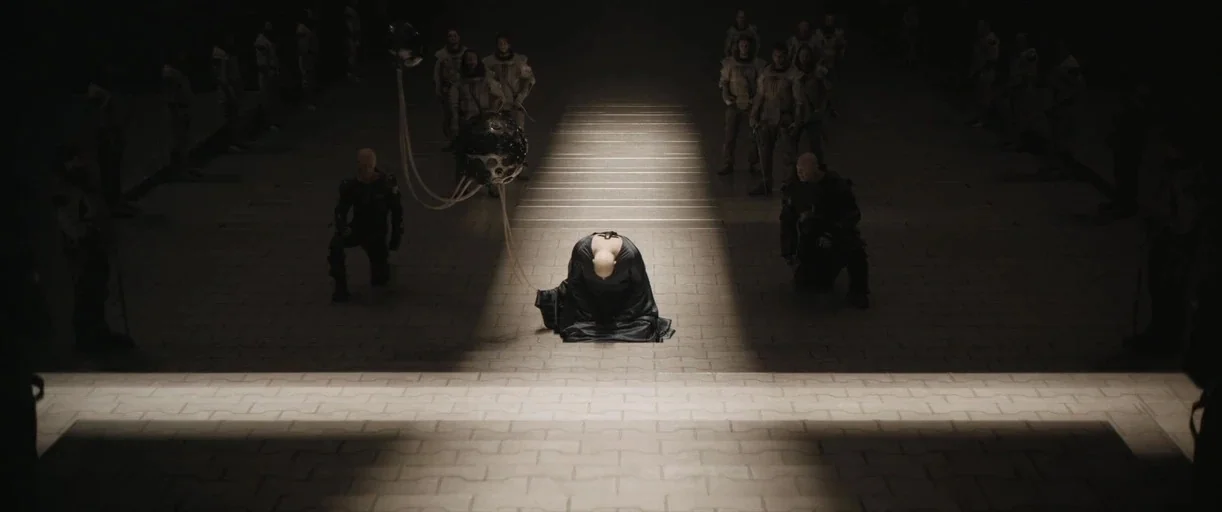 Dune: Part 2 (2024) movie still: wide — A large stone-paved hall is dark except for a singular bright beam of overhead light. A lone figure in dark…