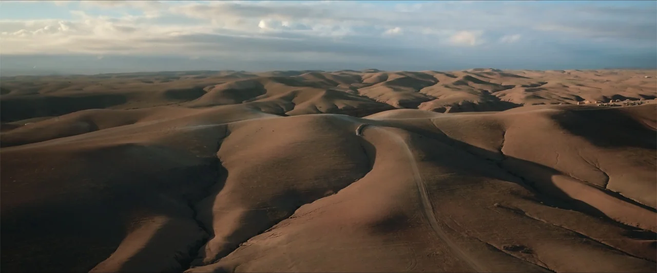 An Endless Wedding (2025) movie still: establishing — An expansive arid desert landscape features undulating hills and a single unpaved dirt road.…