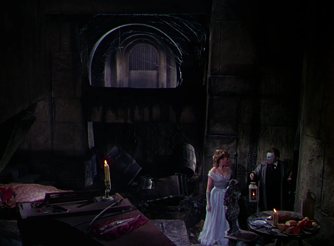 Phantom of the Opera (1943) movie still: wide — A dark, cavernous stone chamber contains a piano and a wooden table with food. A woman in a white…