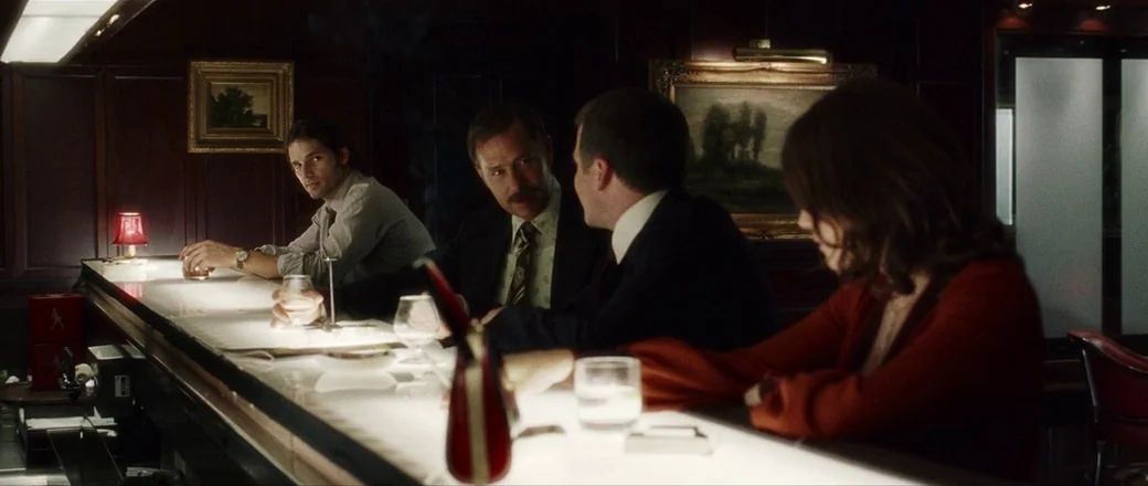 Munich (2005) movie still: medium — A dimly lit upscale bar with dark wood paneling and framed oil paintings on walls. Four people converse while…