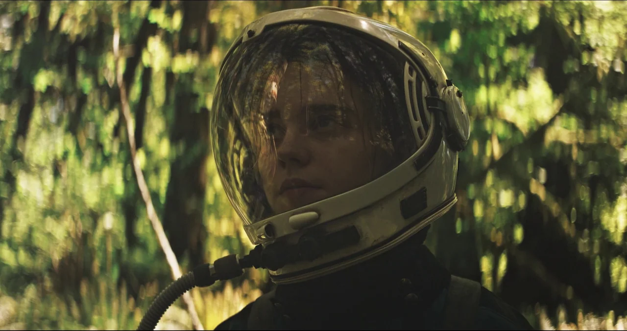 Prospect (2018) movie still: closeup — A dense woodland filled with green foliage surrounds a person wearing a space helmet. The subject stands…