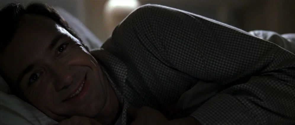 American Beauty (1999) movie still: closeup — A dark, dimly lit indoor setting featuring a person resting on a white pillow. The man is lying down…
