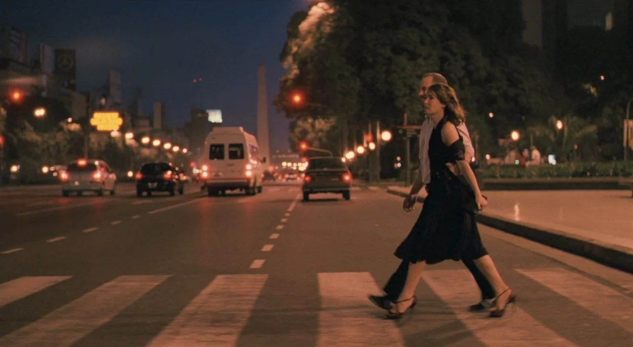 Patagonia (2010) movie still: fullbody — A city crosswalk at night features an obelisk landmark in the distant background. A couple walks across the…