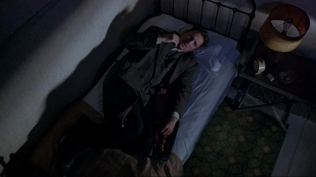 Spider (2002) movie still: medium — A man in a trench coat lies on a simple metal-framed bed at night. A bedside lamp casts a warm glow on the table…