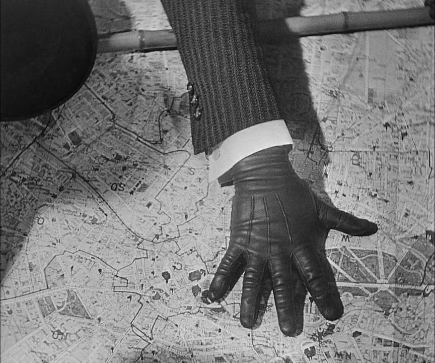 M (1931) movie still: closeup — A close-up view shows a hand in a black leather glove on a map. The map displays a grid of city streets with small…