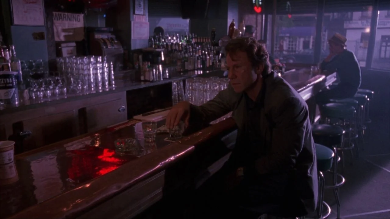 Bad Lieutenant (1992) movie still: medium — A dimly lit neighborhood bar features rows of bottles, stools, and red neon light. A man sits alone at…