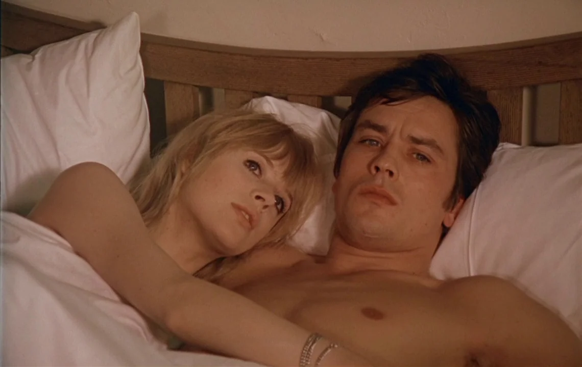 The Girl on a Motorcycle (1968) movie still: closeup — A man and woman lie together in a bed with a wooden headboard. The couple rests under white…
