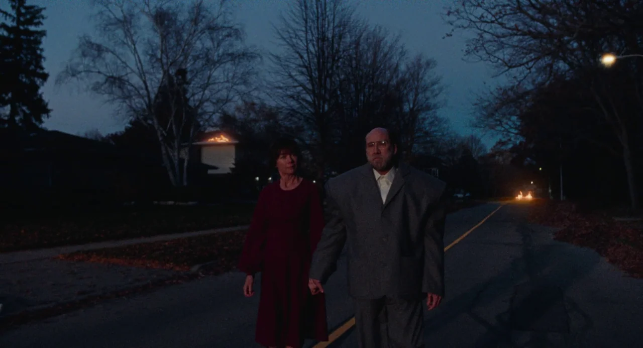 Dream Scenario (2023) movie still: fullbody — A dark residential suburban street at night with leafless trees and a fire burning in distance. An…