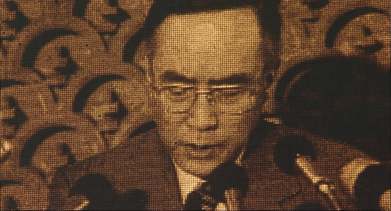 The Return of Godzilla (1984) movie still: closeup — A man speaks into a microphone against a backdrop of stylized graphic logos. The image is…