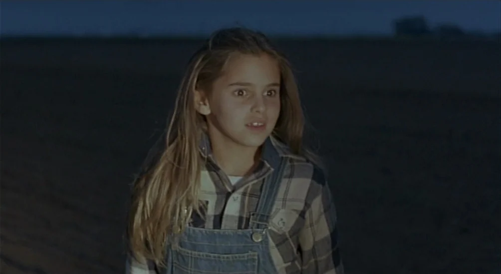 Near Dark (1987) movie still: medium — A dark, empty field extends into the distance under a deep blue night sky. A young girl in plaid stands in…