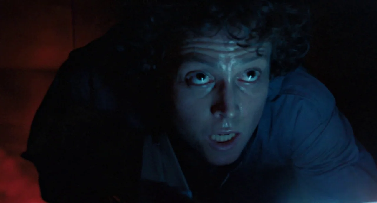 Aliens (1986) movie still: closeup — A dark, cramped metallic environment is lit by harsh contrasting blue and red lights. A woman with curly hair…