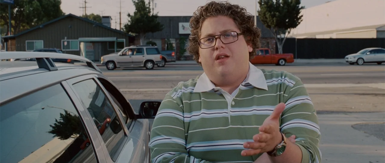 Accepted (2006) movie still: medium — A man stands in a paved lot in front of parked cars. The man wears a striped polo shirt while speaking to…