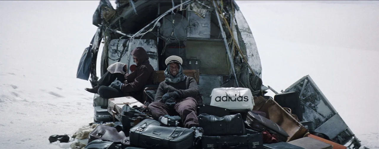 Society of the Snow (2023) movie still: wide — The mangled remains of an airplane fuselage rest atop a vast, featureless snow field. Two young men…