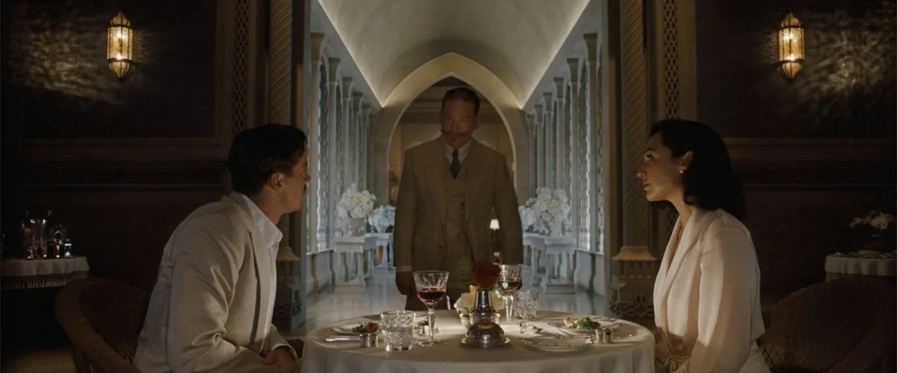 Death on the Nile (2022) movie still: medium — An opulent hotel dining room with arched columns and dim warm lighting. A man stands between two…
