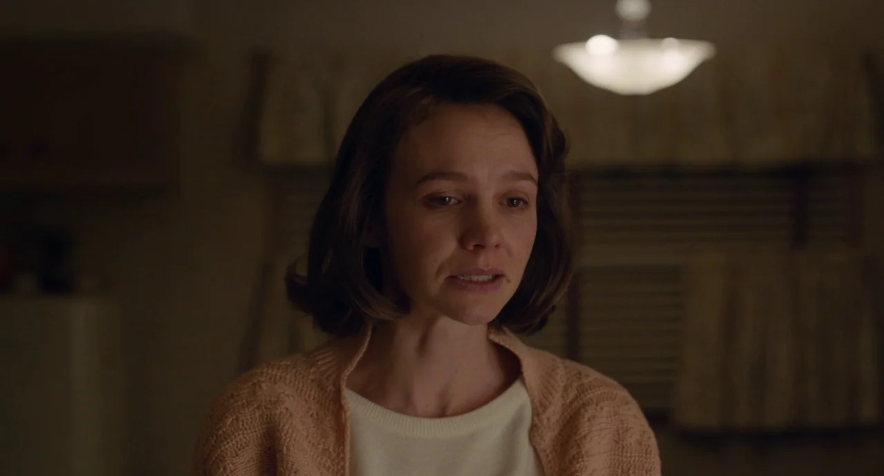 Wildlife (2018) movie still: closeup — A dimly lit indoor space with blurred window blinds in the background. A woman in a knit cardigan speaks…