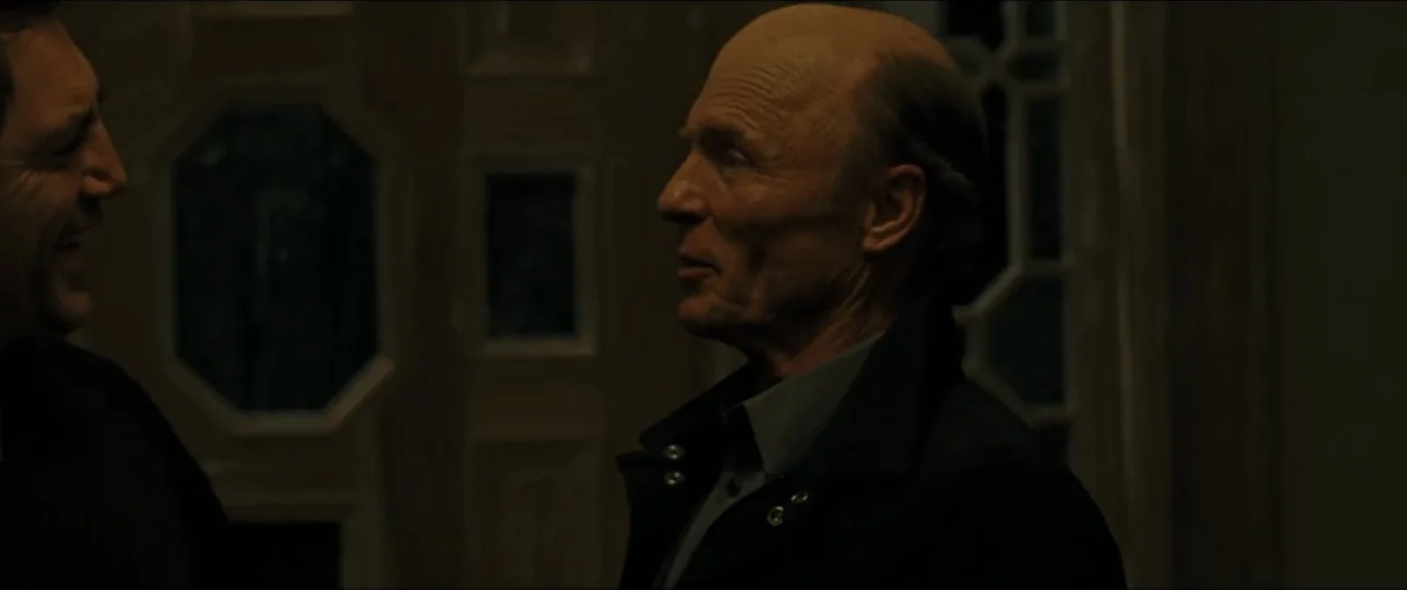 mother! (2017) movie still: medium — A dimly lit indoor corridor with textured wall panels features patterned dark glass insets. Two men engage in a…