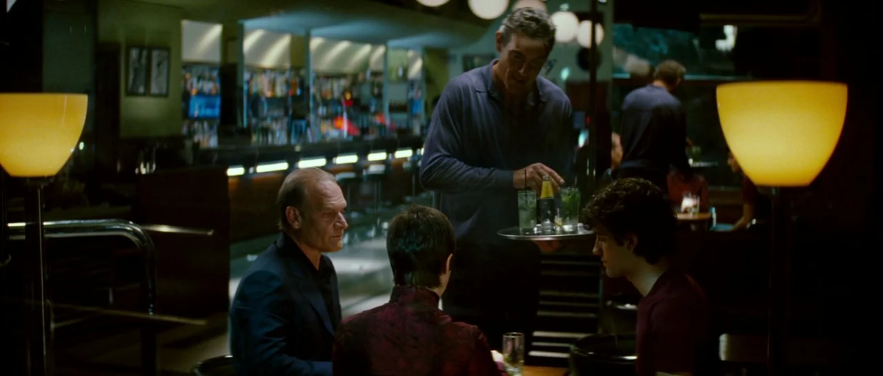 Broken Embraces (2009) movie still: medium — A dimly lit lounge featuring a bar in the back and tables in foreground. A tall man in a polo shirt…