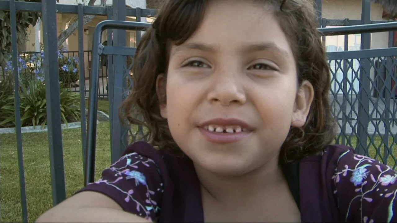 Homeless: The Motel Kids of Orange County (2010) movie still: closeup — A young girl sits outside on a patio with a metal fence backdrop. She faces…