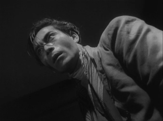 Drunken Angel (1948) movie still: closeup — A man looks upward in a dimly lit, high-contrast interior space. He appears to be reacting to something…