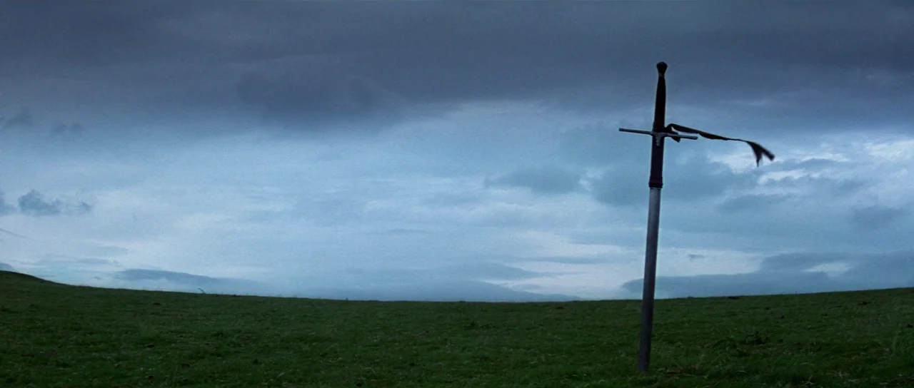 Braveheart (1995) movie still: wide — A wide, grassy field sits under a dense, overcast gray-blue sky. A lone longsword is stuck vertically into the…