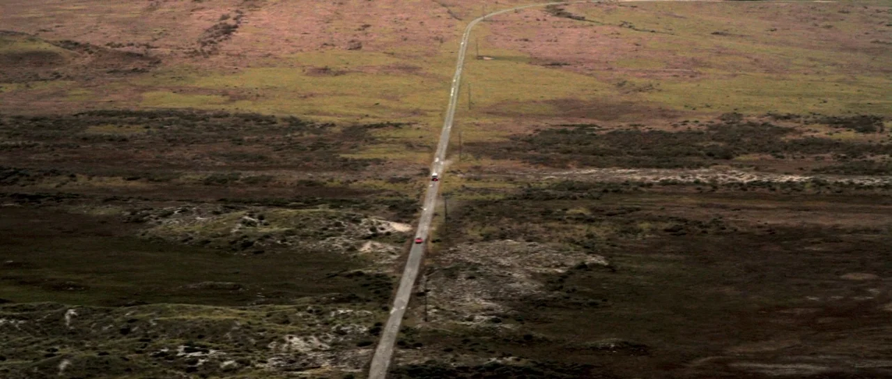 I Melt With You (2011) movie still: establishing — An aerial view shows a narrow, paved road cutting through a wide, textured moorland. Two small…