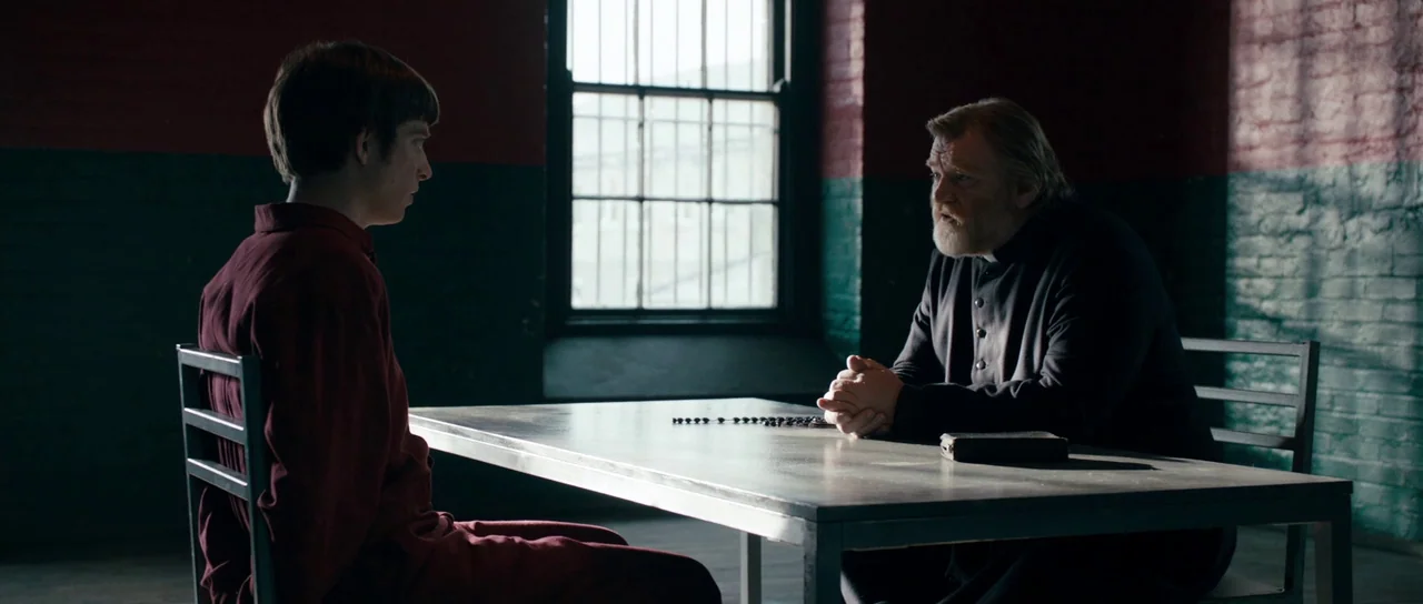 Calvary (2014) movie still: medium — A dark visitation room with painted brick walls and a metal table. The priest faces a young man in prison…
