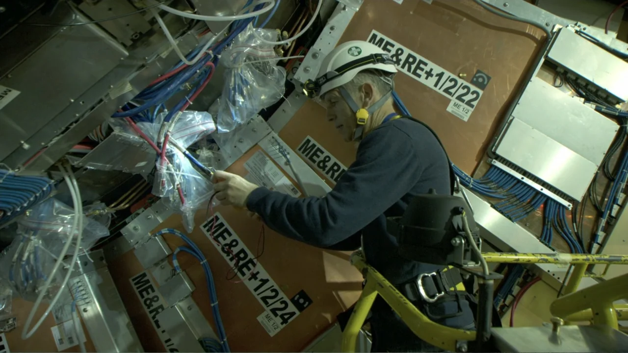 CERN (2013) movie still: medium — A complex technical enclosure filled with labeled panels and tangled electrical cabling. A technician wearing a…