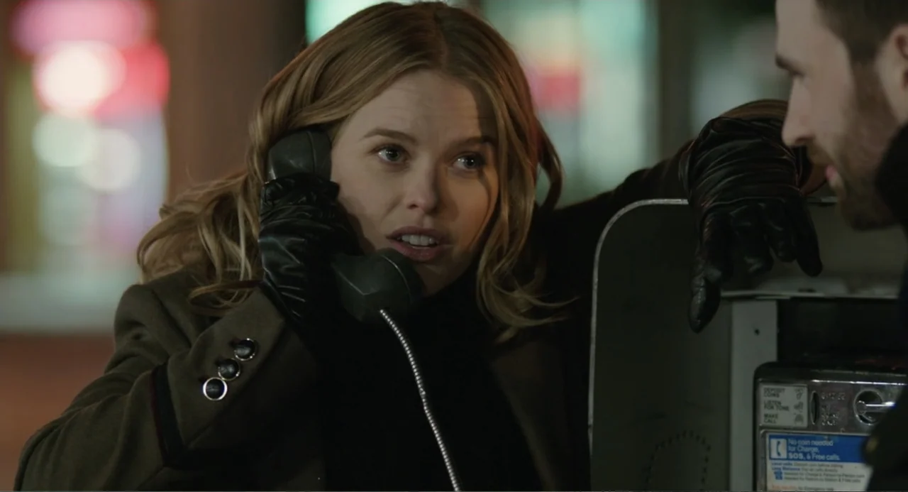 Before We Go (2014) movie still: medium — A dark outdoor urban environment features a lit metal payphone kiosk at night. A woman in an olive coat…