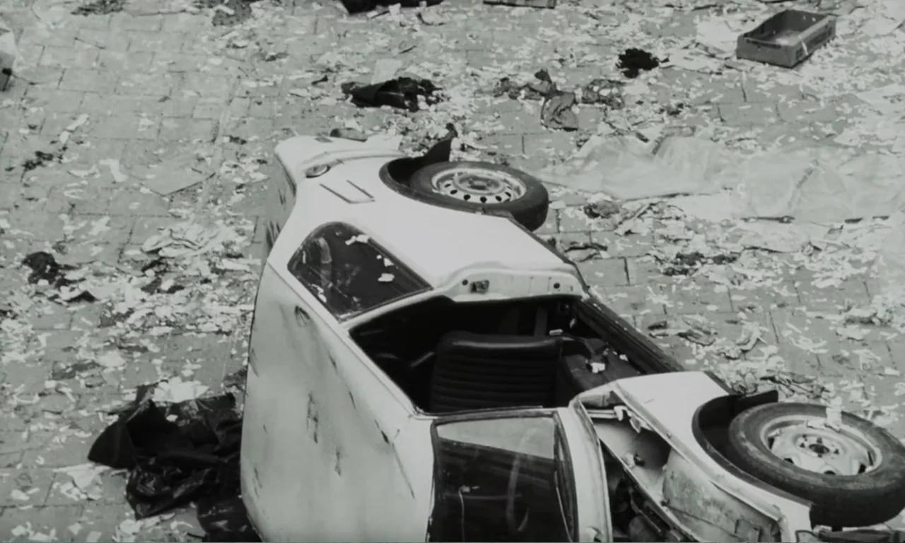 The Sacrifice (2015) movie still: wide — A paved city square is littered with white papers and scattered debris. An overturned white automobile lies…