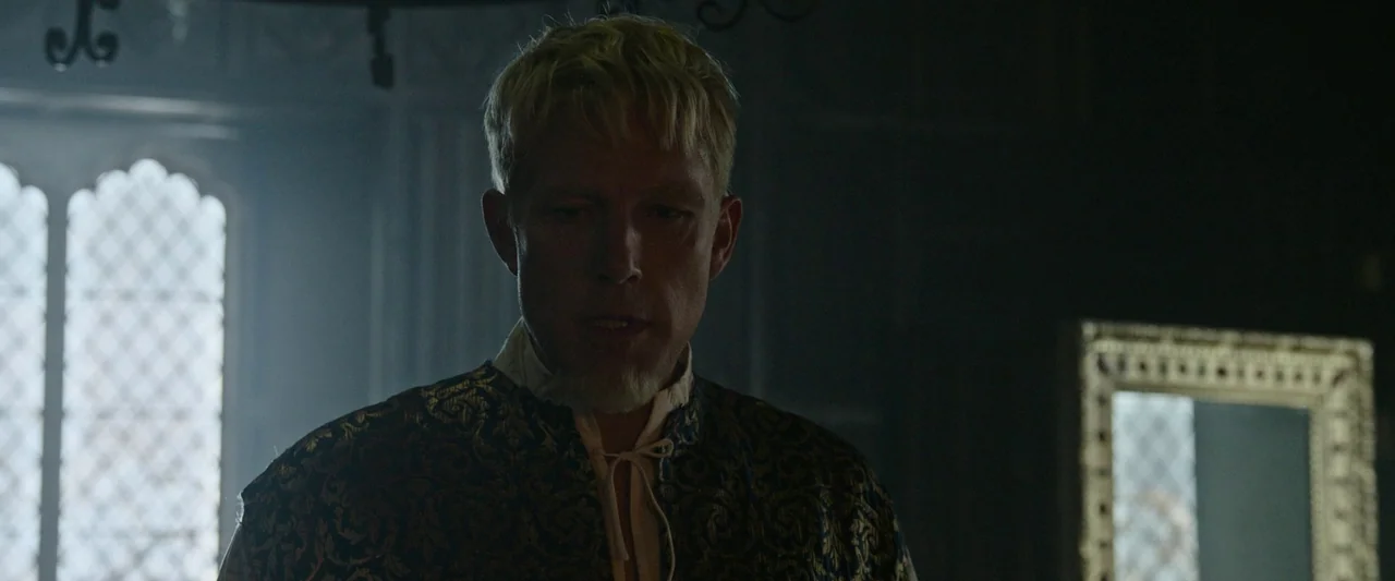 The Last Duel (2021) movie still: medium — A dark, dimly lit room features a Gothic-style window and an ornate gilded mirror. A blonde man wearing…