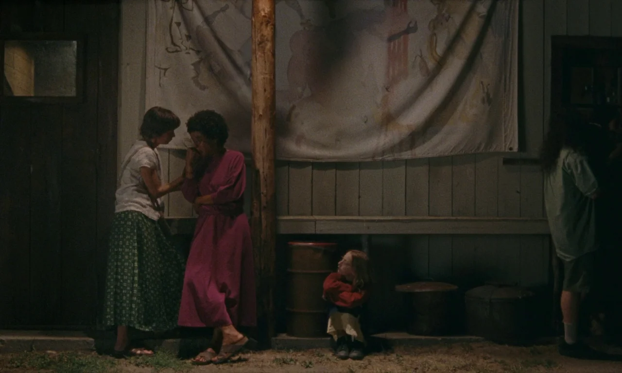 Janet Planet (2023) movie still: fullbody — A wooden porch at night features a fabric banner pinned against the siding wall. Two women stand talking…