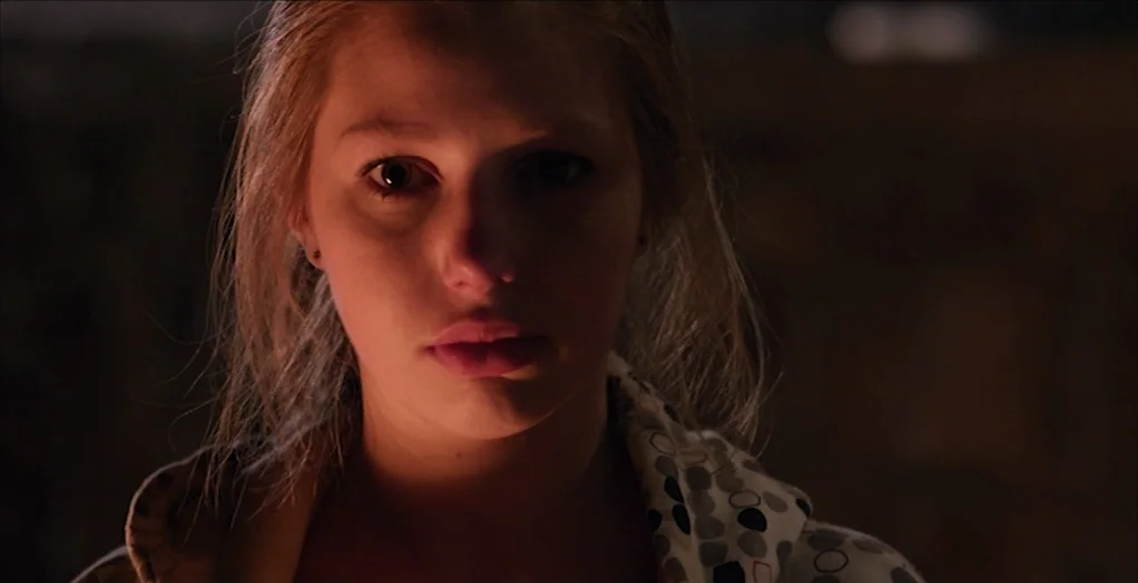 Dollhouse (2012) movie still: closeup — A dark space with a person illuminated against a dimly lit backdrop. The subject remains motionless while…