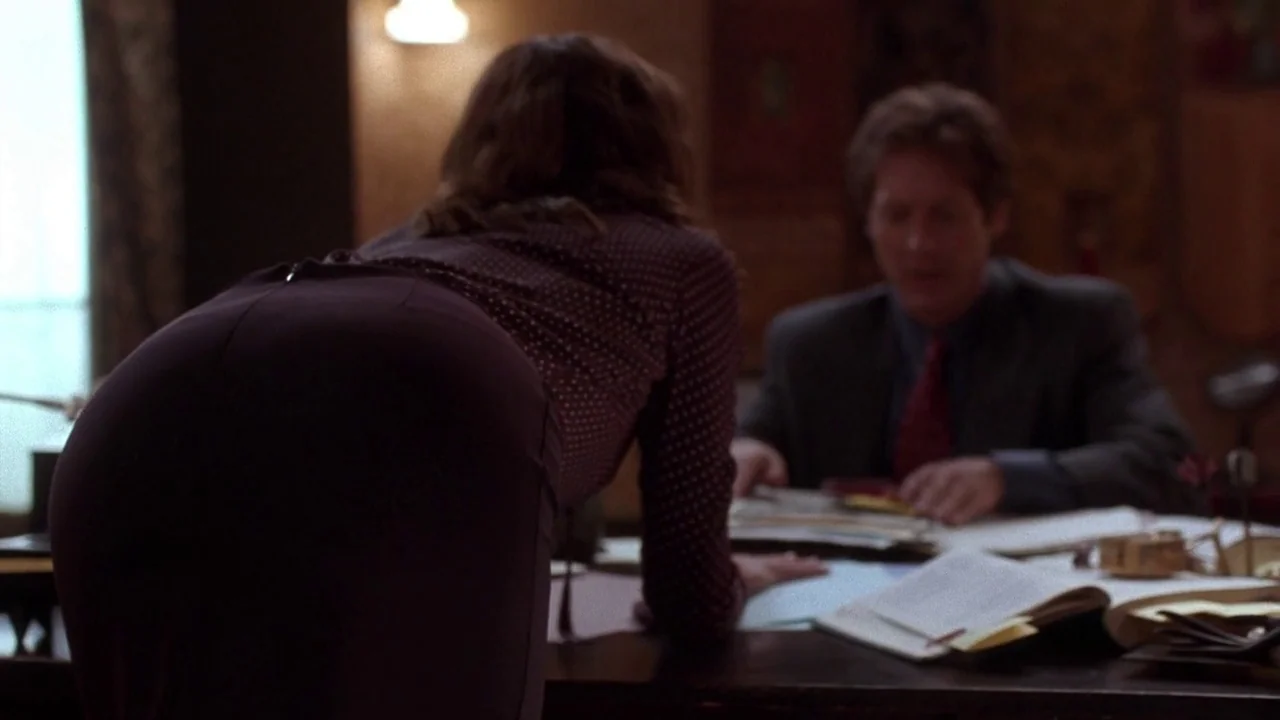 Secretary movie still: medium — A cluttered wood desk occupies the foreground and center of a dimly lit room. A woman leans forward with her back to…