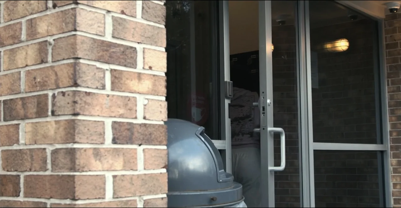 The Devil Is Busy (2024) movie still: medium — A brown brick building facade features a glass door partially open to an interior. A person in a grey…