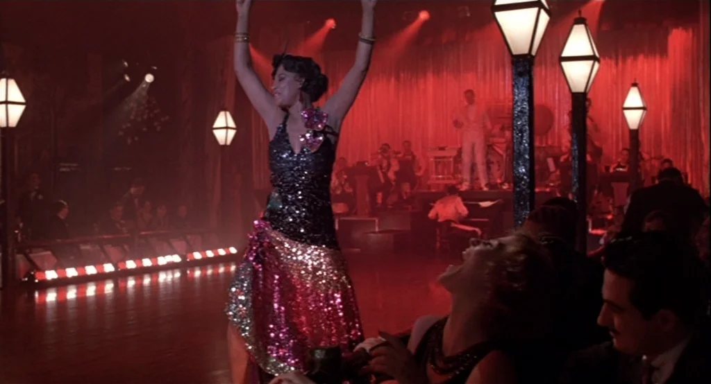 The Cotton Club (1984) movie still: fullbody — A dimly lit nightclub features a wooden floor and a bandstand in the background. A woman in a…