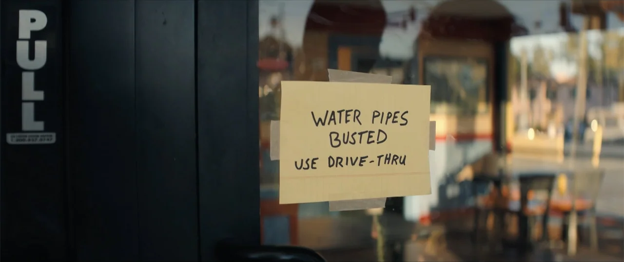 One of Them Days (2025) movie still: closeup — A yellow paper note is taped to the exterior glass door of a restaurant. The sign conveys a message…