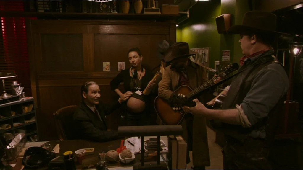 A Prairie Home Companion movie still: medium — A dark, wood-paneled office contains desk clutter and musical instruments. A man sits at a desk while…