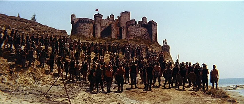 Erik The Conqueror (1961) movie still: wide — A stone castle sits atop a rugged coastal cliff overlooking a sandy beach below. A large group of…