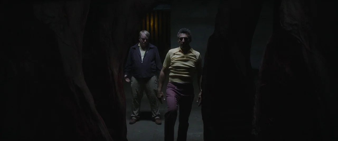 God’s Pocket (2014) movie still: fullbody — Two men walk through a dark, narrow corridor carved from rough, textured stone surfaces. They move…