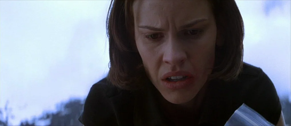 Insomnia (2002) movie still: closeup — An outdoor area with blurred foliage visible in the out-of-focus background. A young woman looks down with an…