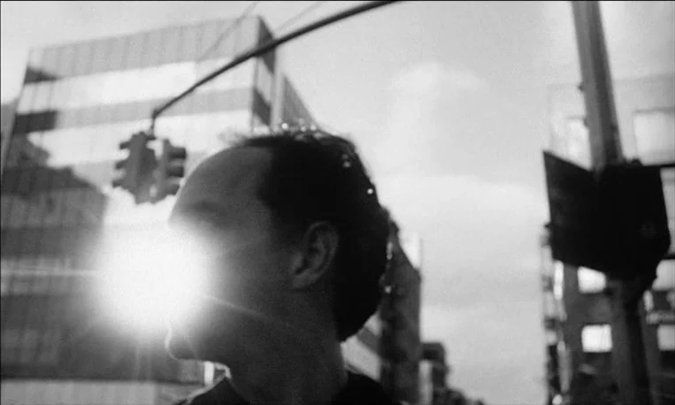 Pi (1998) movie still: closeup — A man stands on a blurred city sidewalk with high-rise buildings in the distance. A bright sun flare completely…