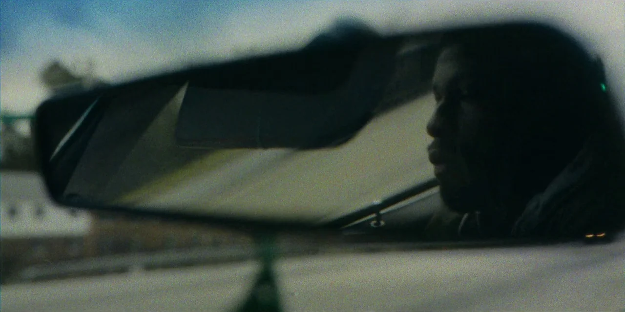 They Cloned Tyrone (2023) movie still: closeup — The interior of a vehicle seen reflected in the rearview mirror. A man is visible inside the mirror…