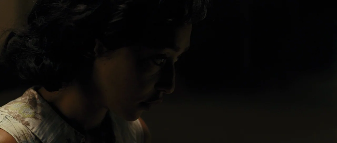 Loving (2016) movie still: closeup — A woman's face is illuminated against a dark background in a dimly lit room. She looks downward with a neutral…