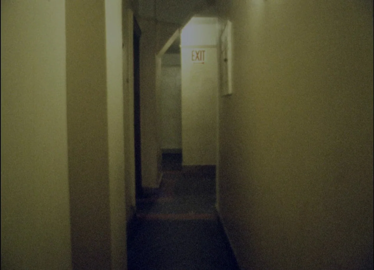 Hotel Monterey (1973) movie still: establishing — A narrow, dimly lit apartment hallway features beige walls and a dark floor. A red exit sign is…