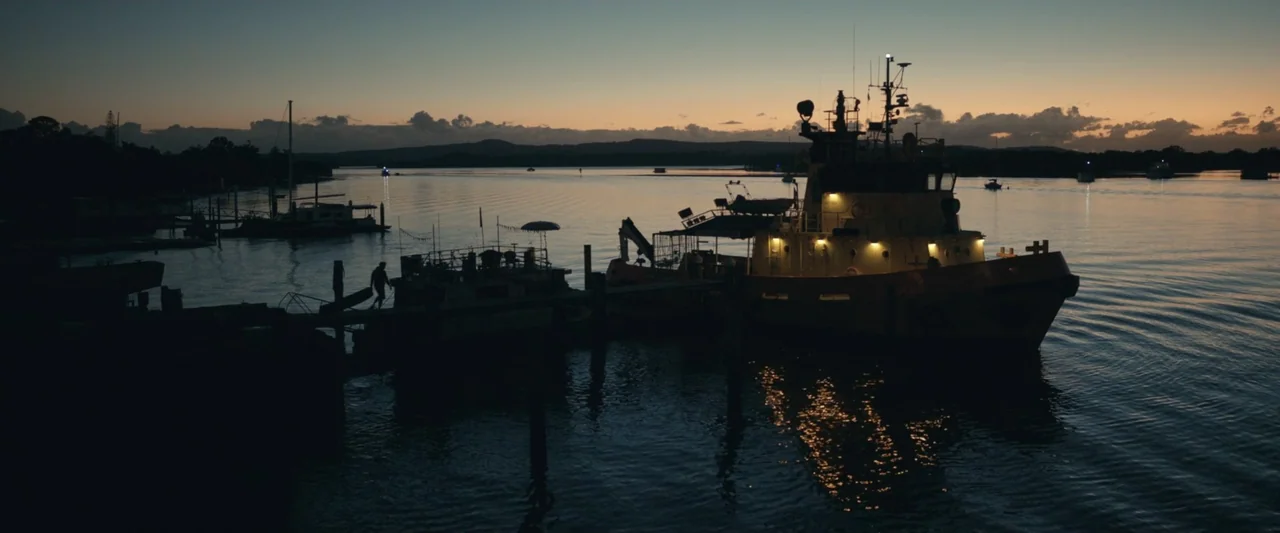 Dangerous Animals (2025) movie still: wide — A harbor at dusk features a tugboat docked alongside a wooden pier. The calm water reflects the soft…