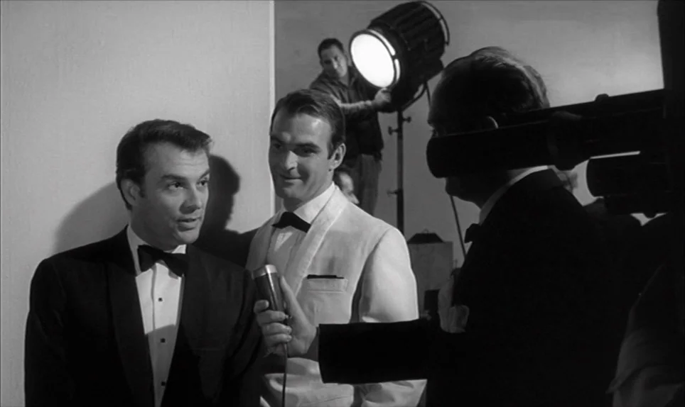 Eva (1962) movie still: medium — A professional television studio setting featuring plain walls and a bright overhead production light. Two men in…