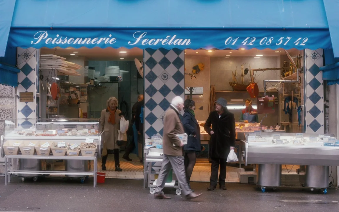 Let The Sunshine In (2017) movie still: wide — A fish store storefront with blue awnings features a diamond-patterned tile facade. Several people…