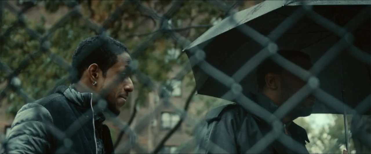 Exposed (2016) movie still: medium — An outdoor area featuring a prominent chain-link fence obscuring a view of a building. Two men stand close…