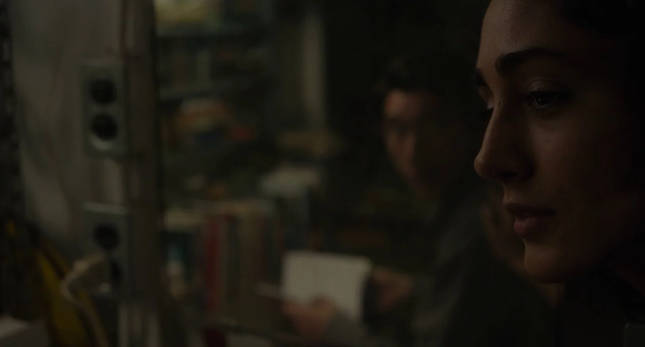 Paterson (2016) movie still: closeup — A dark, cluttered workspace contains electrical outlets and stacked items in the background. A woman in…